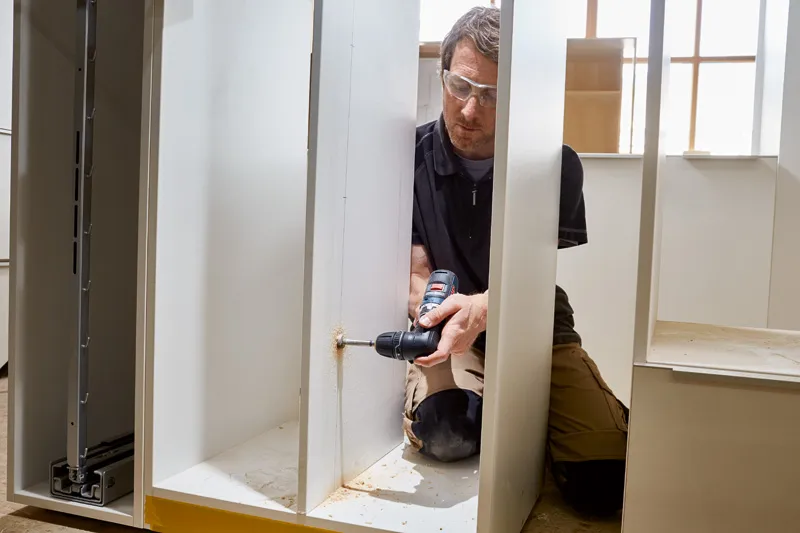 En person iført sikkerhedsudstyr borer ind i et skabspanel og danner træstøv.